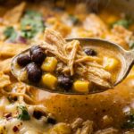 Ultra close-up macro of comforting creamy chicken tortilla soup spoon lifting tender shredded halal chicken, black beans, sweet corn, and crisp tortilla strips
