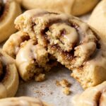 Ultra close-up macro of bakery-style cinnamon roll cookie broken in half showing tender crumb, gooey cinnamon sugar spiral swirl, and cream cheese icing drizzle
