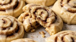 Ultra close-up macro of bakery-style cinnamon roll cookie broken in half showing tender crumb, gooey cinnamon sugar spiral swirl, and cream cheese icing drizzle