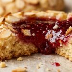 Ultra close-up macro of golden buttery shortbread thumbprint cookie rolled in sliced almonds with glistening ruby raspberry jam center